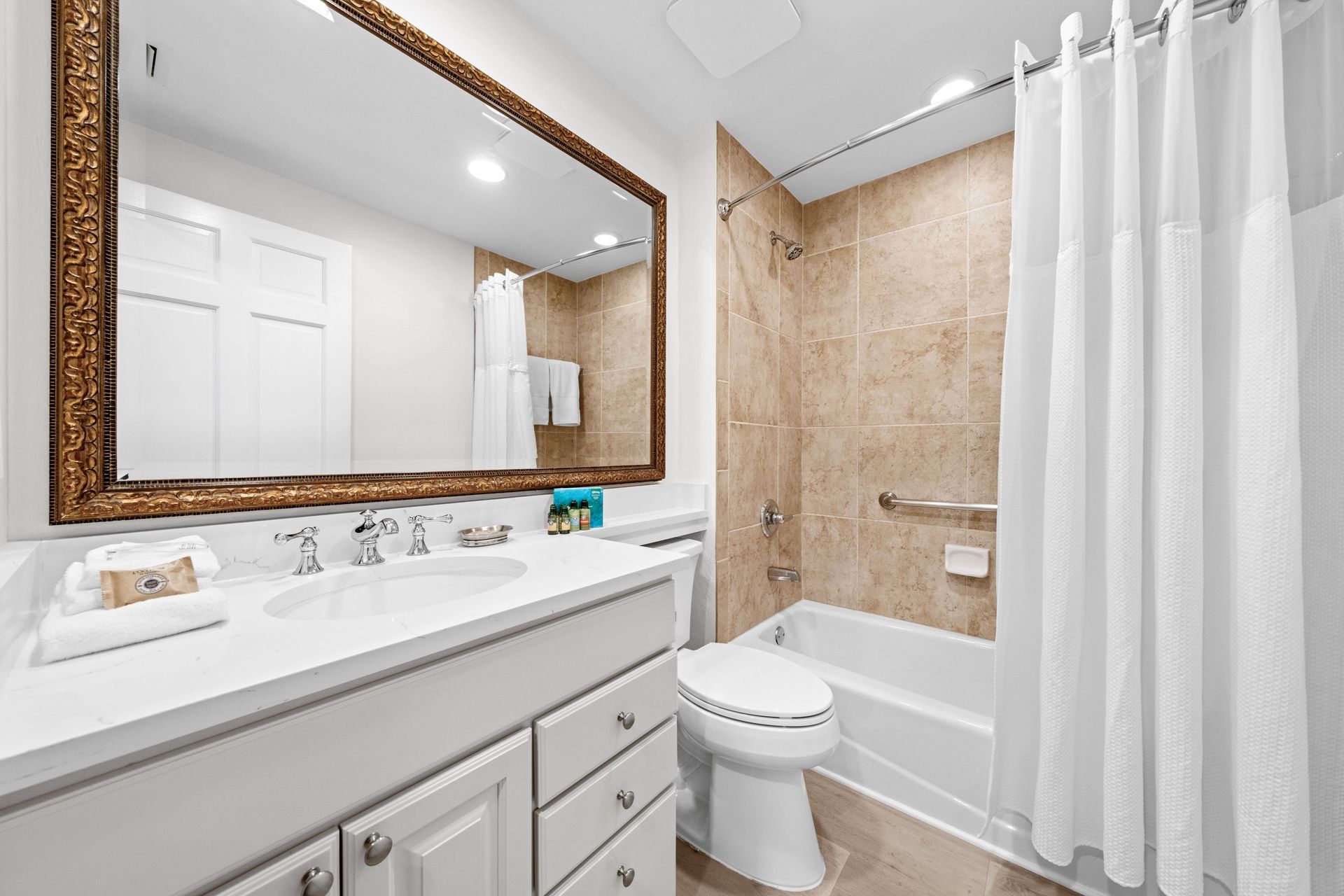 Bright bathroom with vanity, large mirror, toilet, bathtub, beige tile, and white shower curtain.