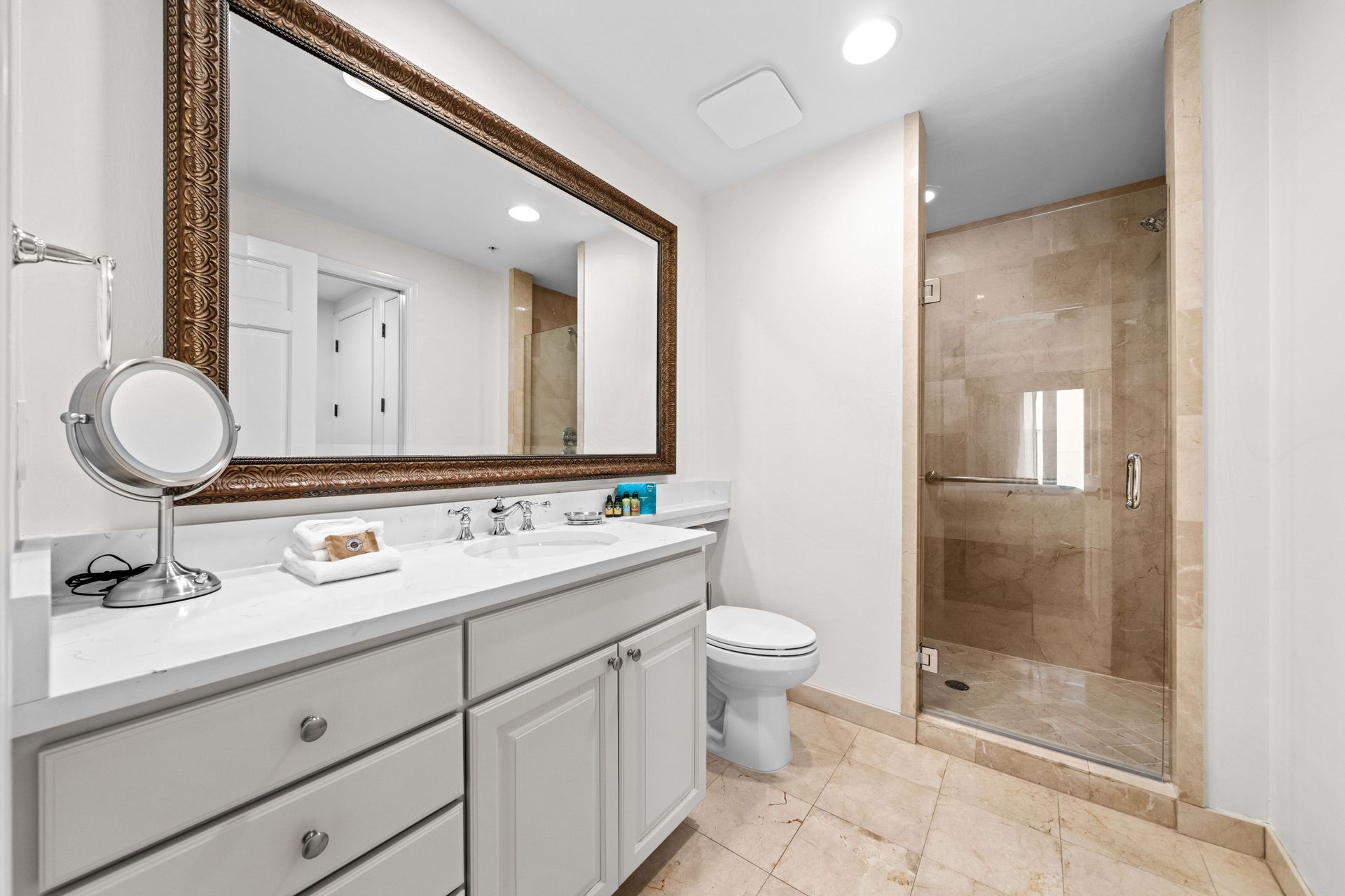 Bright modern bathroom with double vanity, large mirror, toilet, and glass-enclosed shower with beige tile.