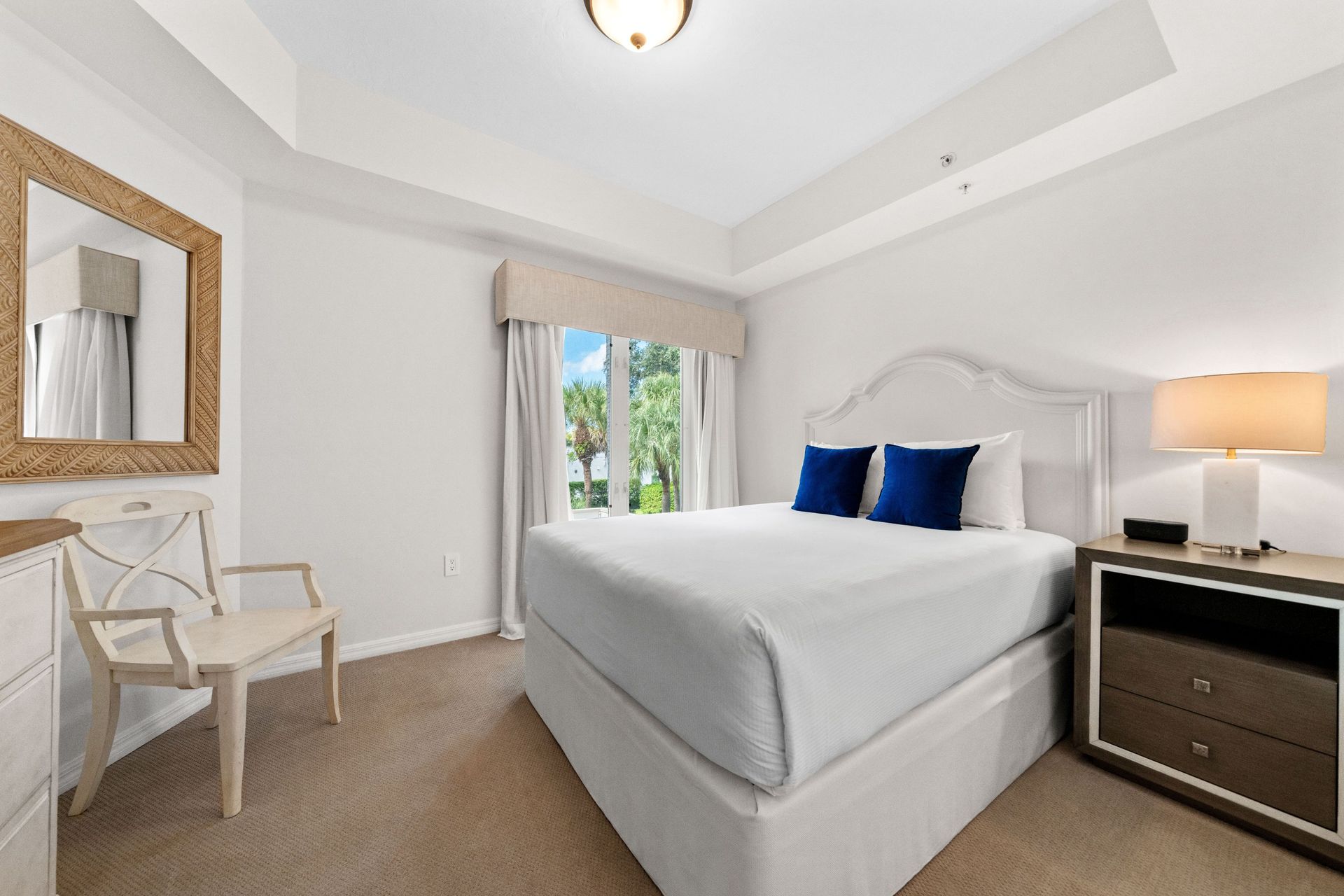 Bright bedroom with white bed, blue pillows, bedside lamp, and chair by a window