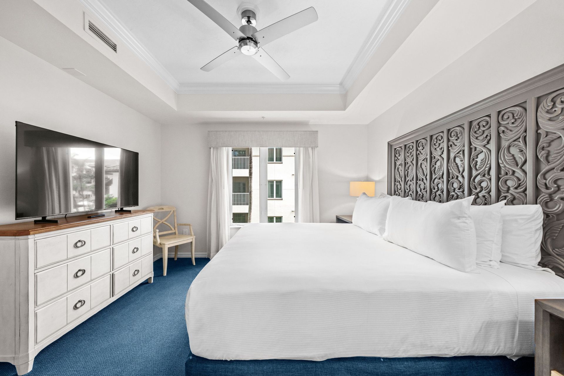 Bright hotel room with king bed, blue carpet, white dresser, TV, and balcony doors