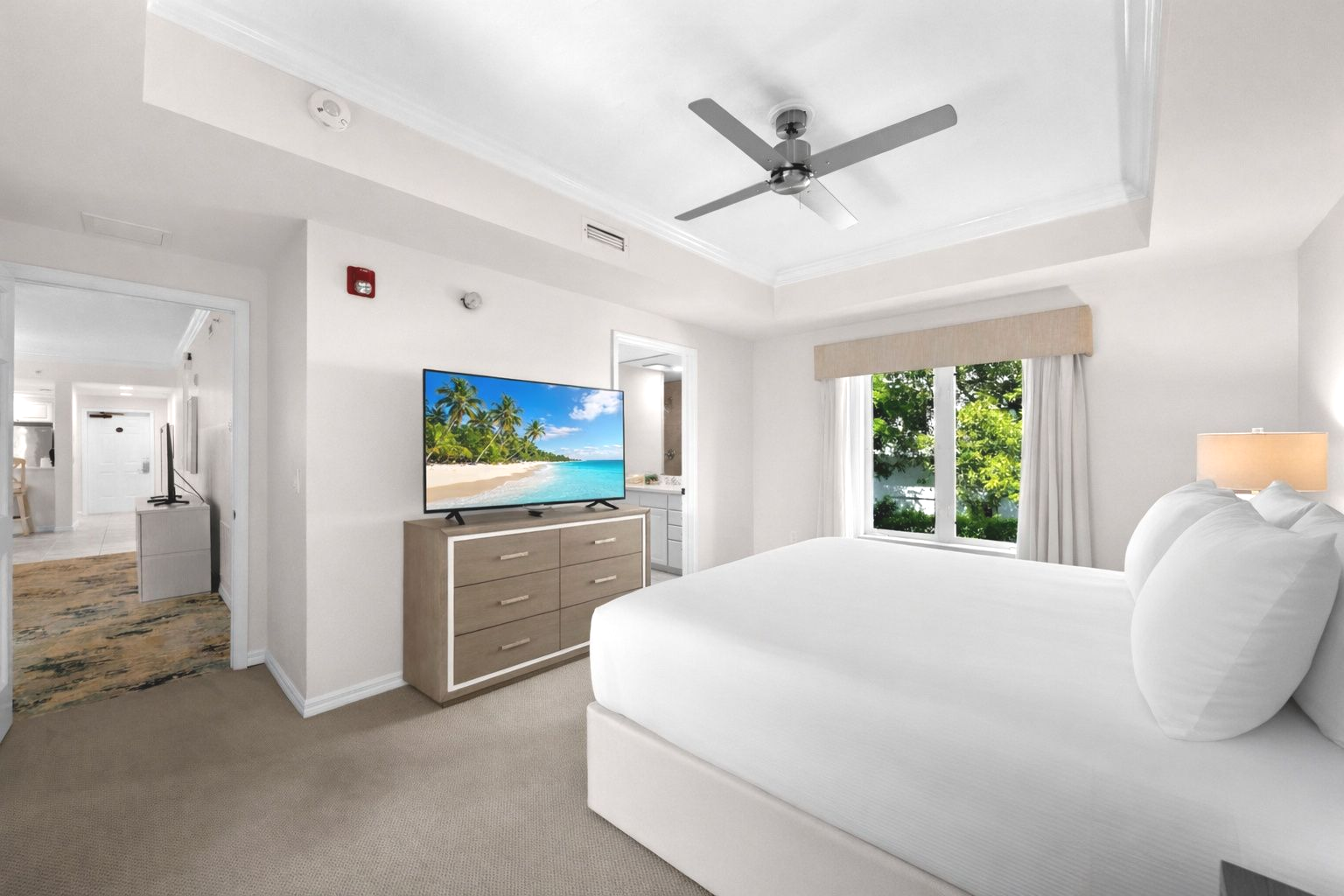 Bright modern bedroom with a white bed, dresser, ceiling fan, and sliding doors to greenery outside.