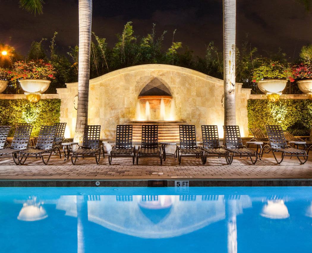 Nighttime resort pool with lit lounge chairs, palm trees, and a stone fountain wall reflected in the water