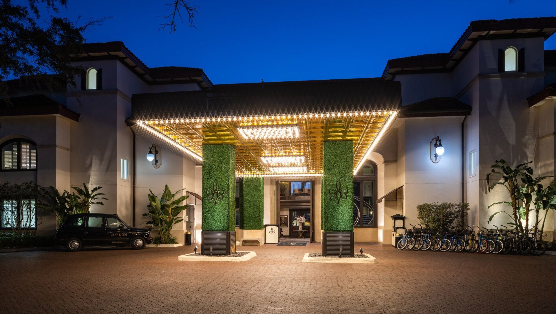 A hotel entrance with a lit canopy, plants, and a car and bicycles parked outside.