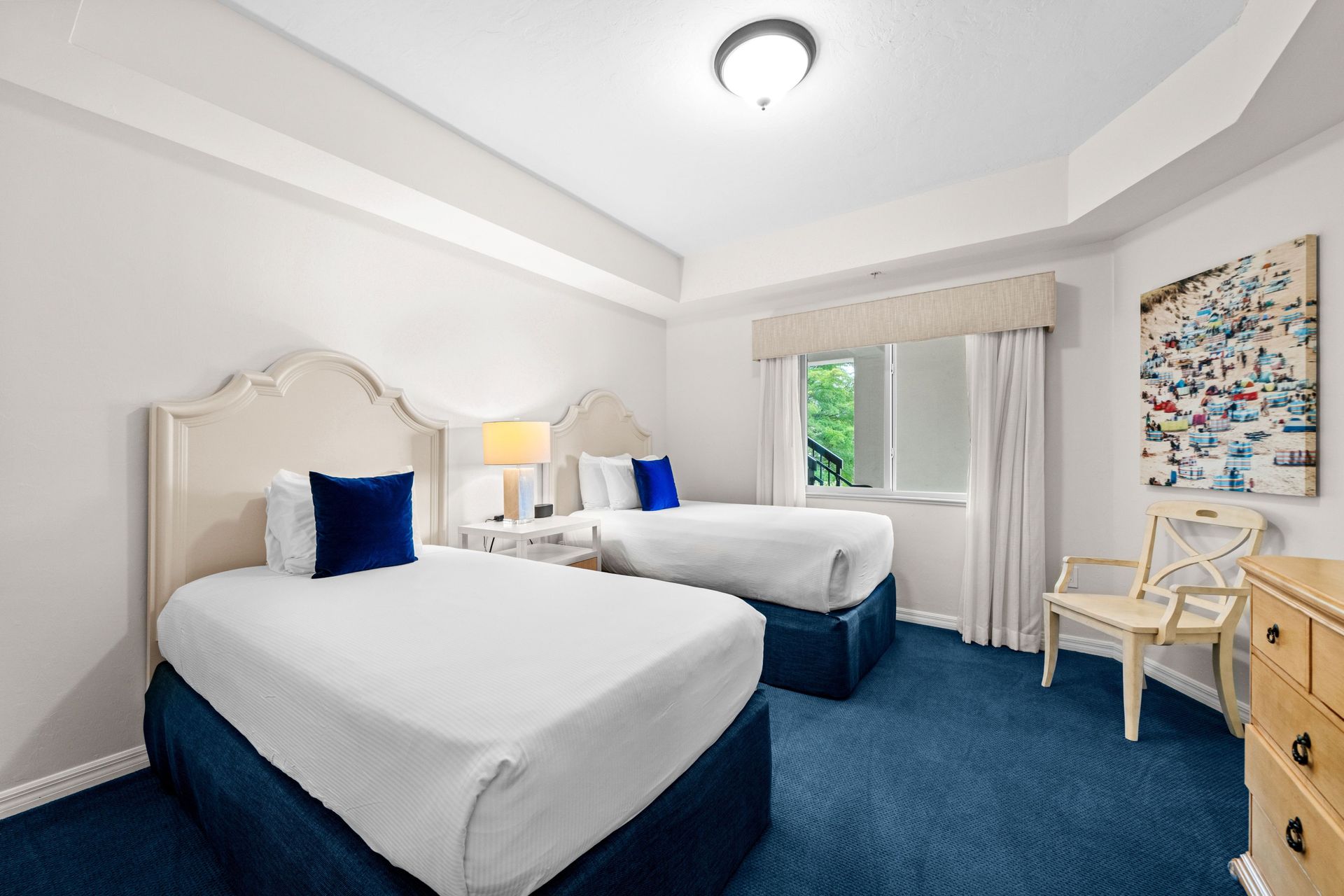 Bright bedroom with two twin beds, blue carpet, a window, and a small desk by the wall