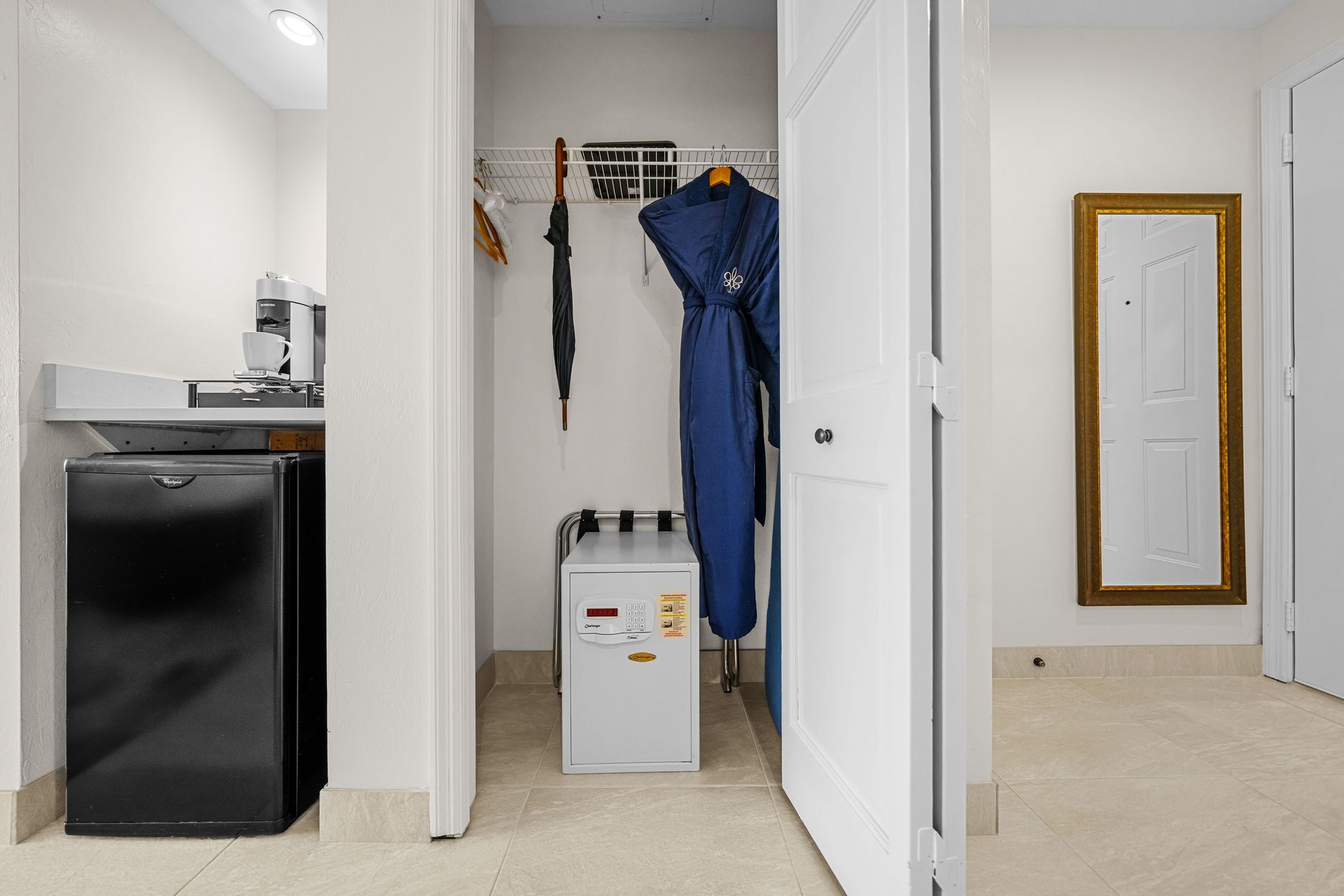Small apartment entryway with a mini fridge, hanging coat and umbrella, and a wall mirror.