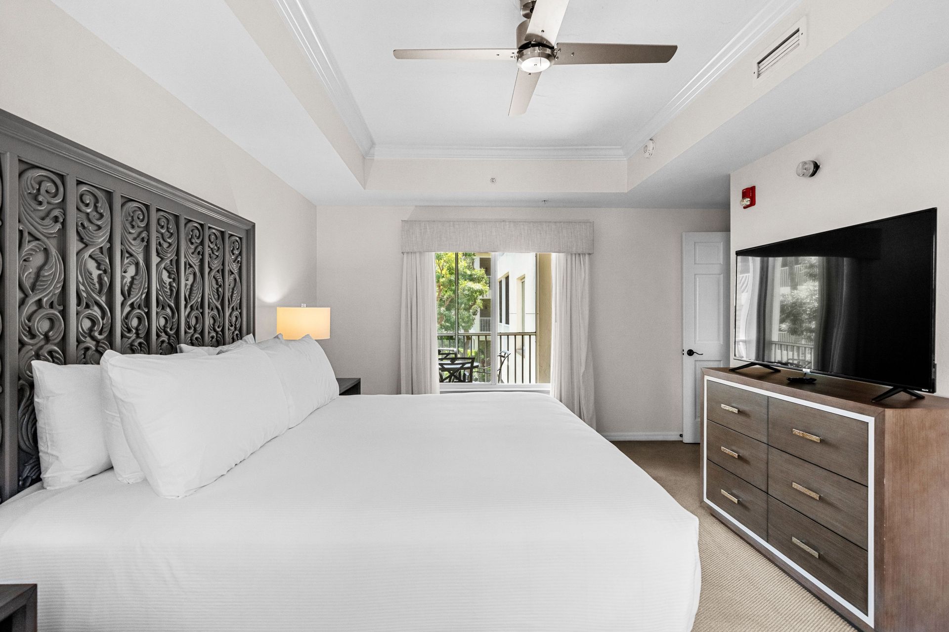 Bright white bedroom with a large bed, ornate headboard, TV, dresser, and balcony doors.