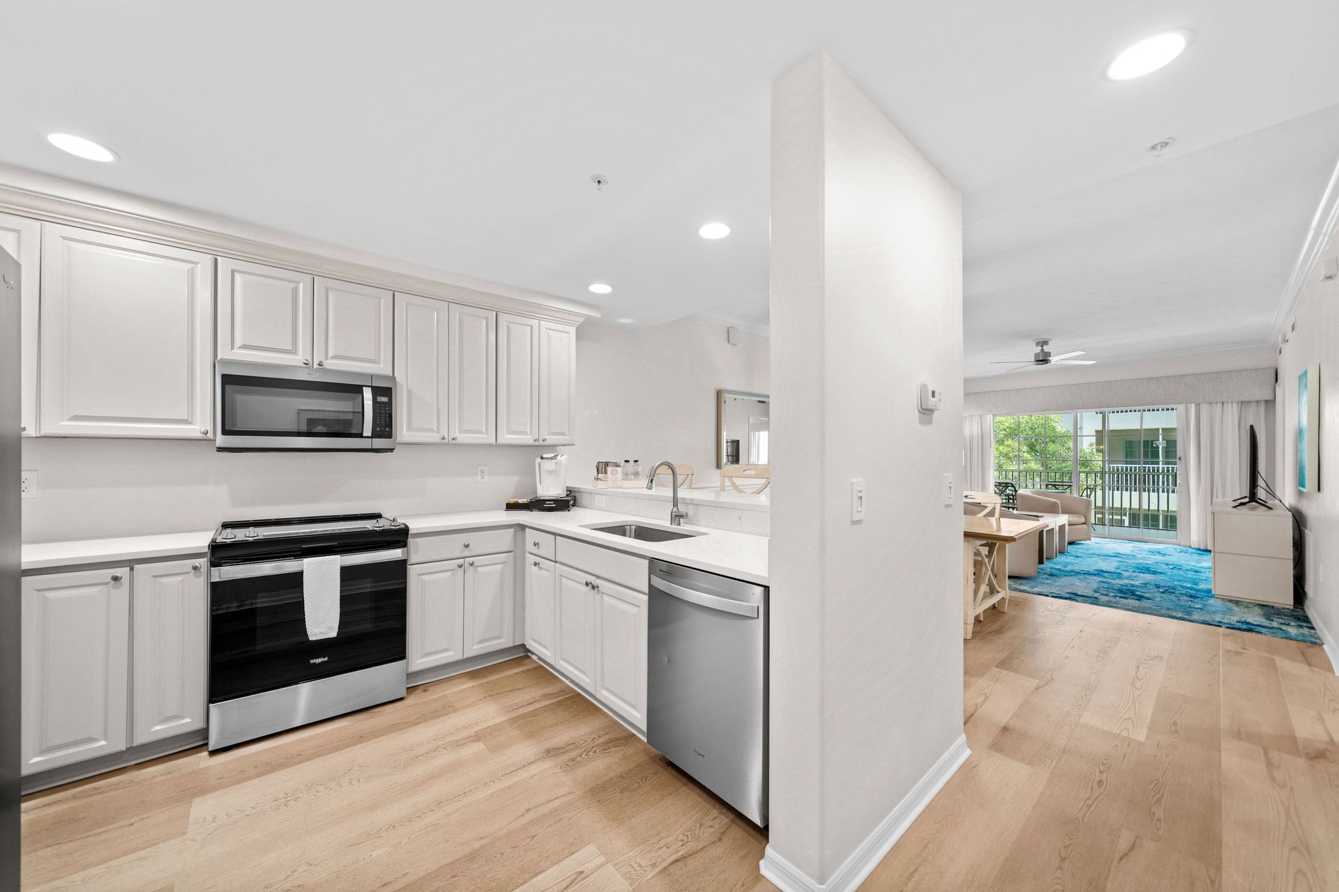 Bright white kitchen with stainless appliances, open to a living area with blue rug and natural light