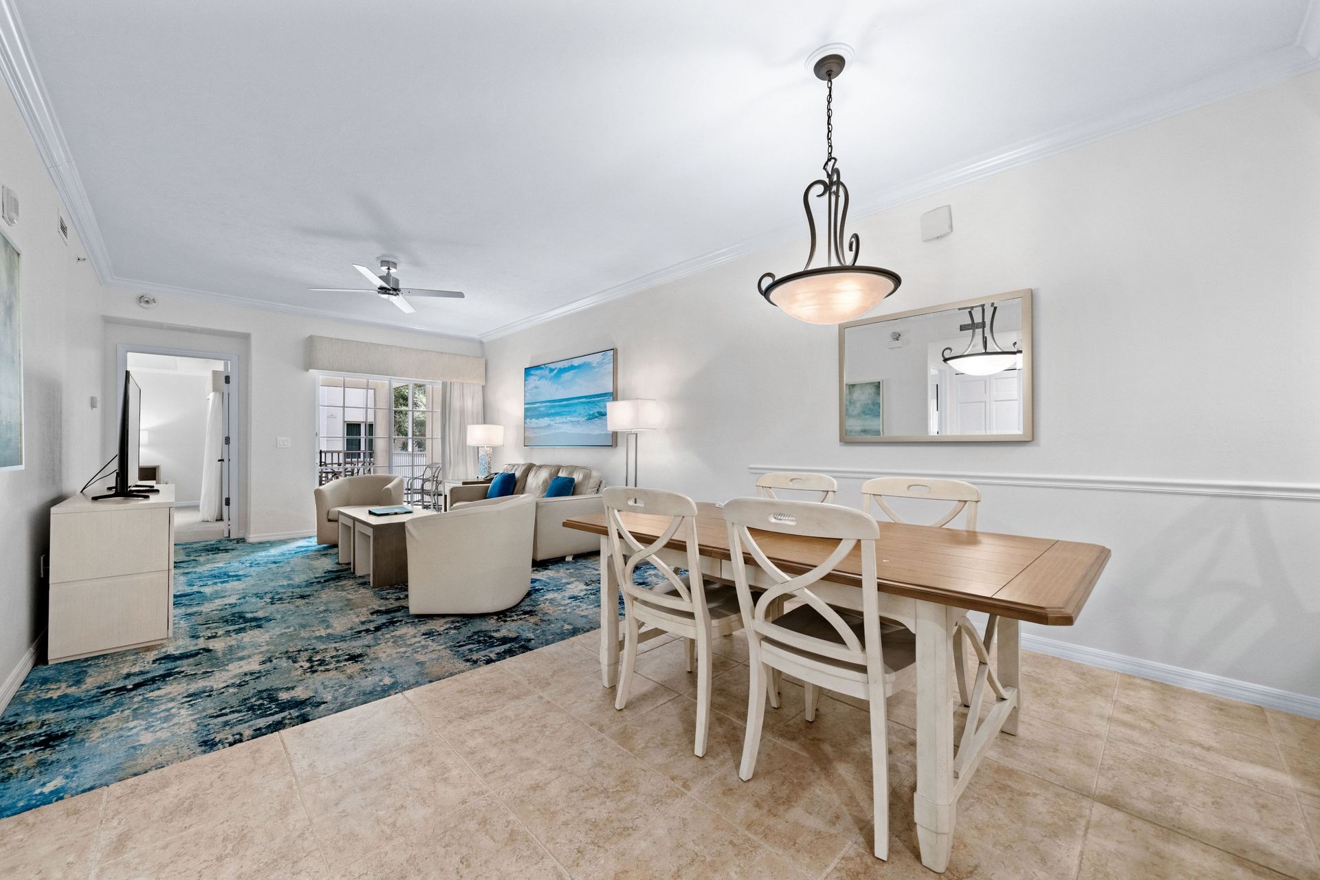 Bright open dining and living room with beige carpet, blue rug, white chairs, and modern chandelier