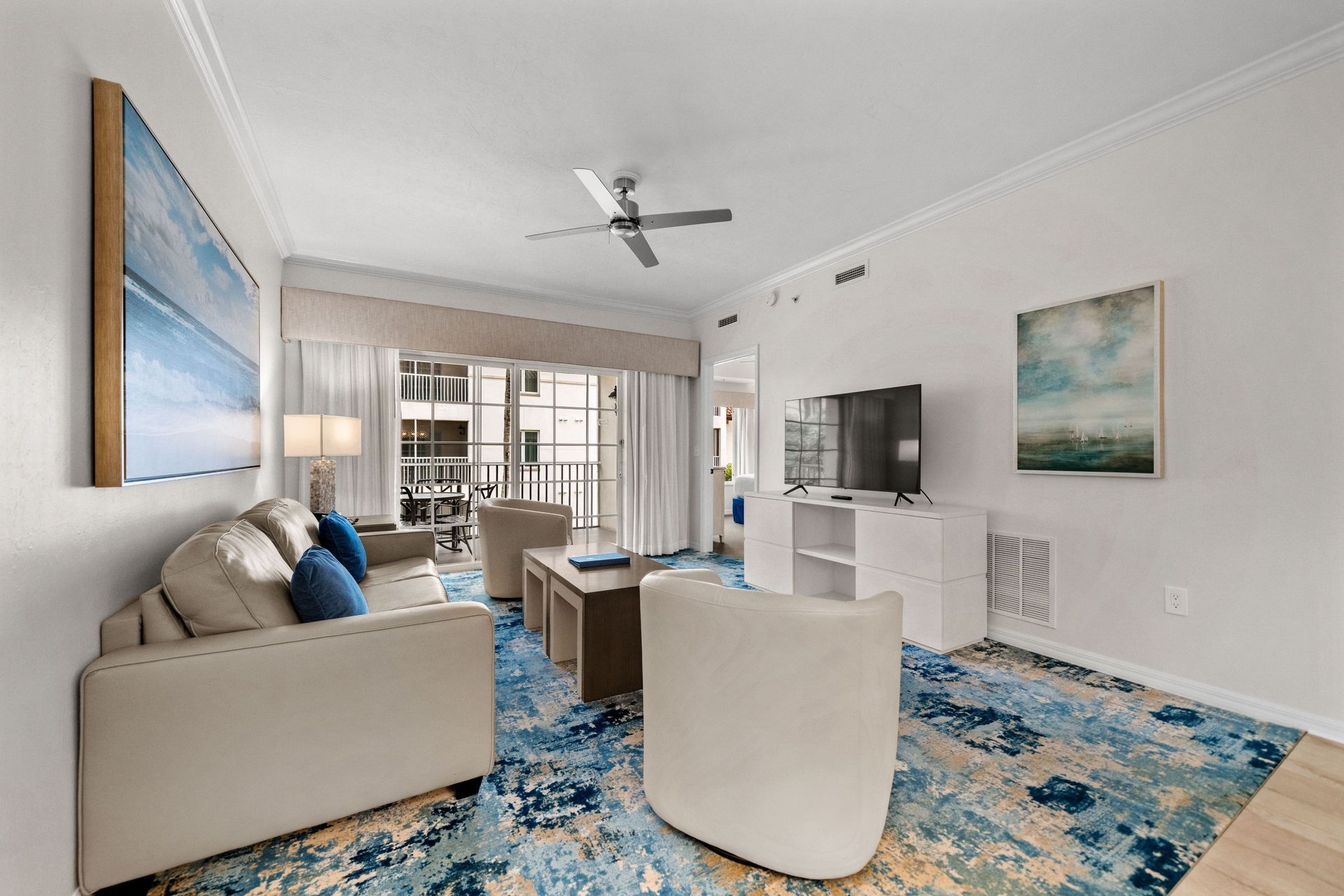 Modern living room with blue patterned rug, beige sofa, white chairs, wall TV, and ocean artwork