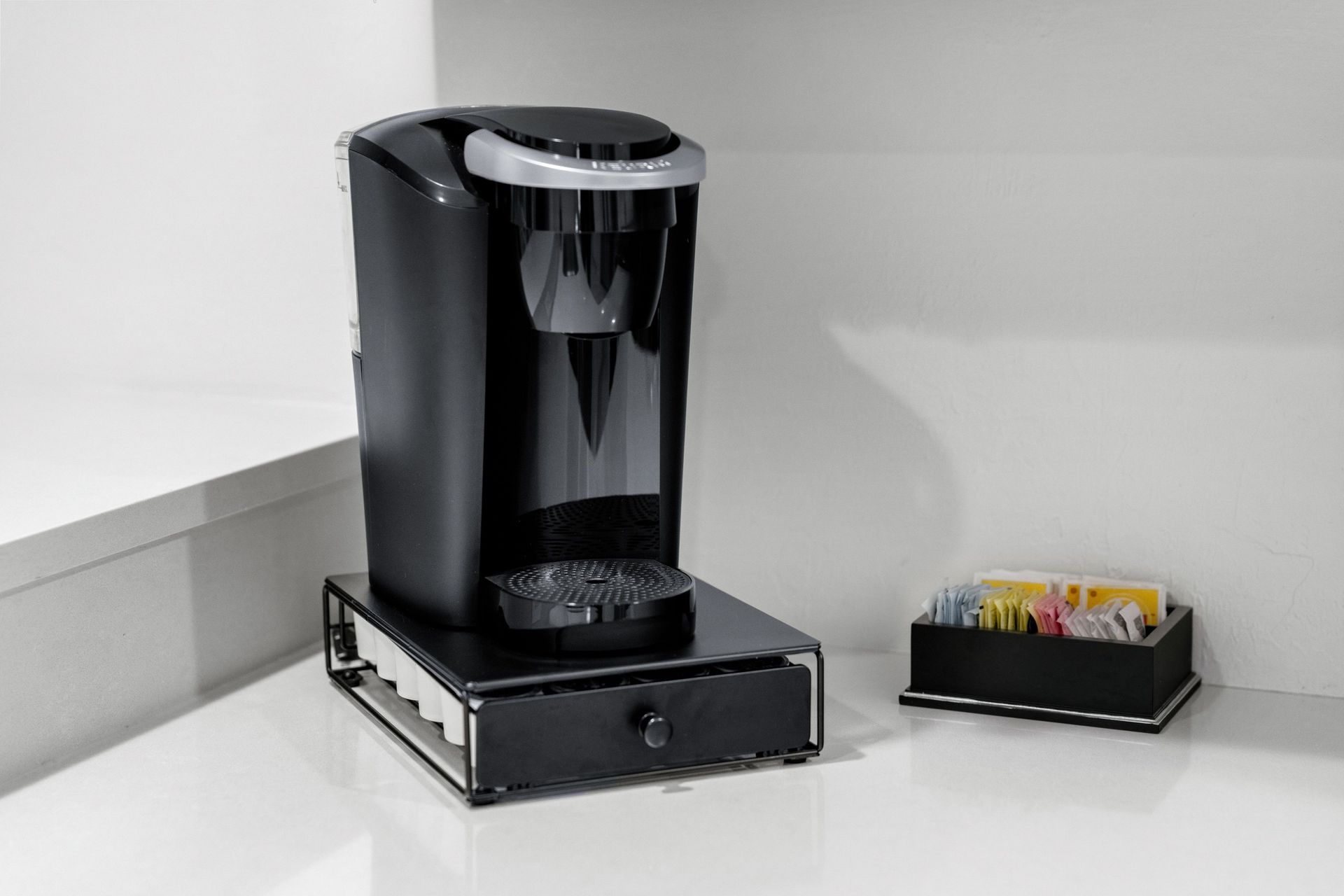 Black coffee maker on a white counter beside a small tray of tea packets