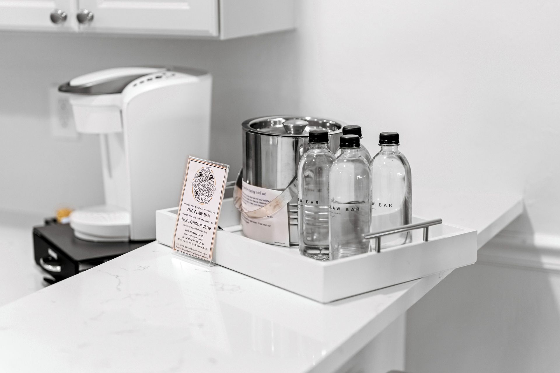 White kitchen counter with coffee maker, glass jars, water bottles, and a tray of tea packets