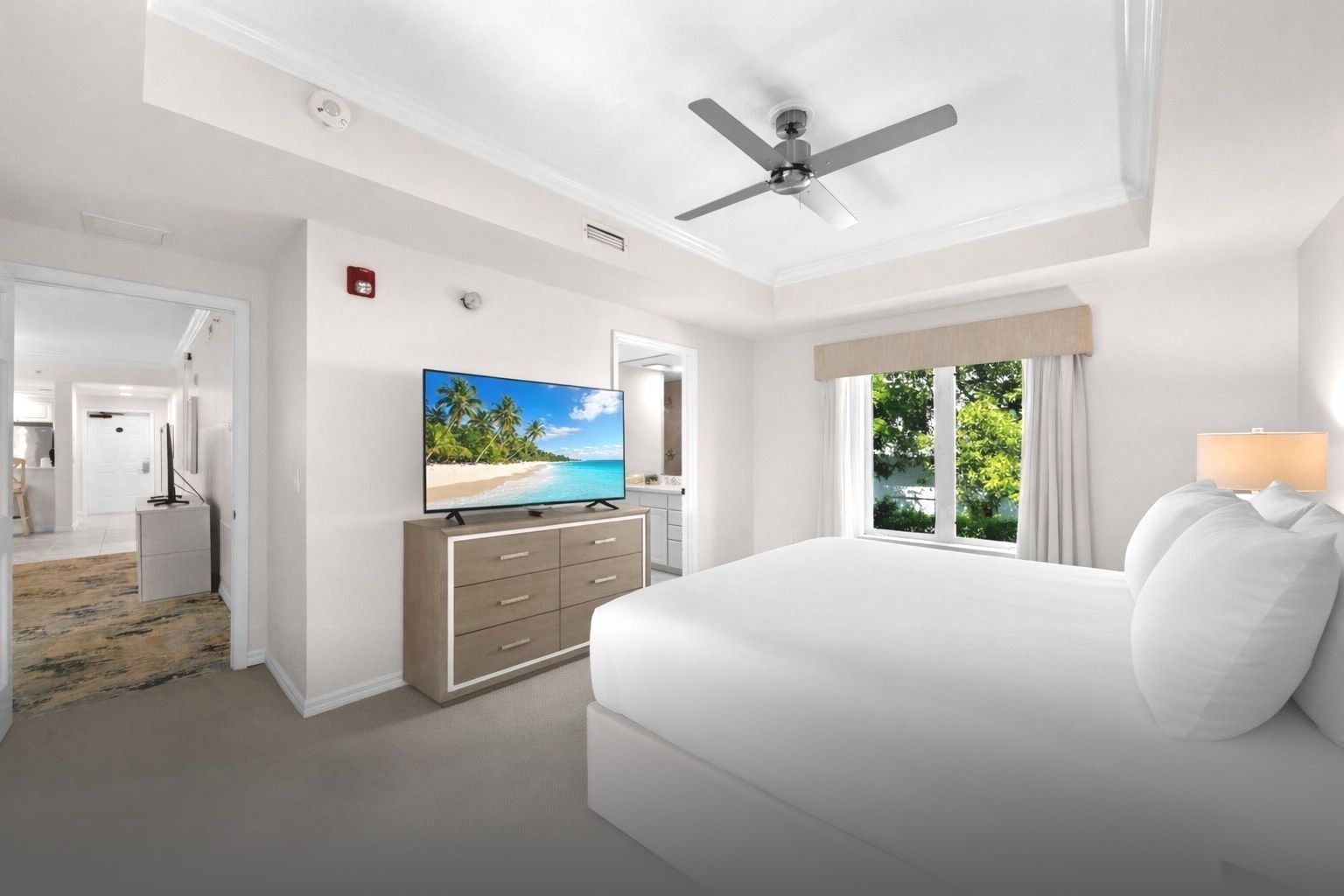 Bright white bedroom with a king bed, dresser and TV, ceiling fan, and large windows overlooking greenery