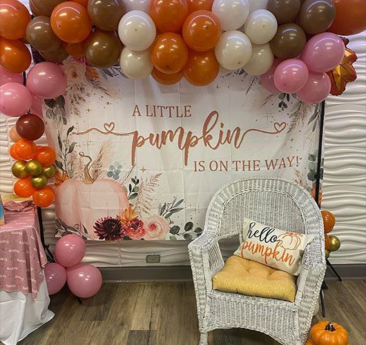 Pumpkin-themed baby shower setup. Includes balloons, banner with chair and pillow that reads 