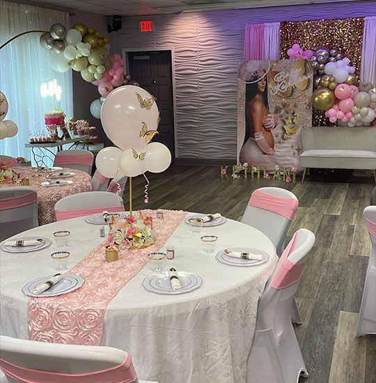 Pink and gold themed event space with decorated tables, balloons, and a backdrop; likely a baby shower.