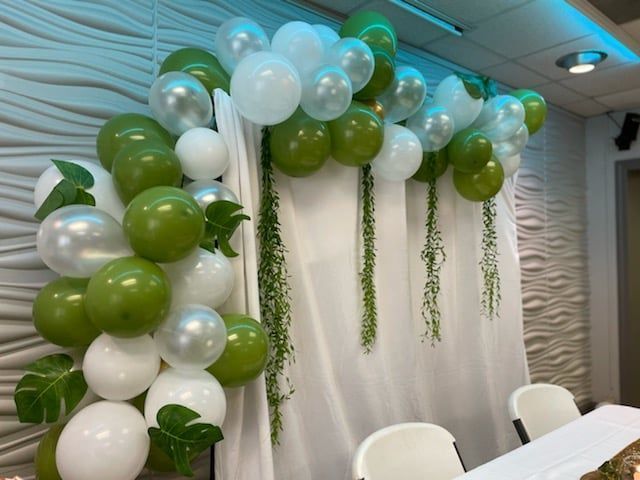 Green, white, and silver balloon arch with greenery against white curtain backdrop.