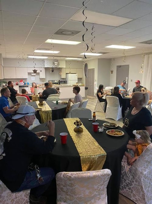 Party in a brightly lit room with guests seated at tables; black tablecloths with gold runners; small kitchen in the background.