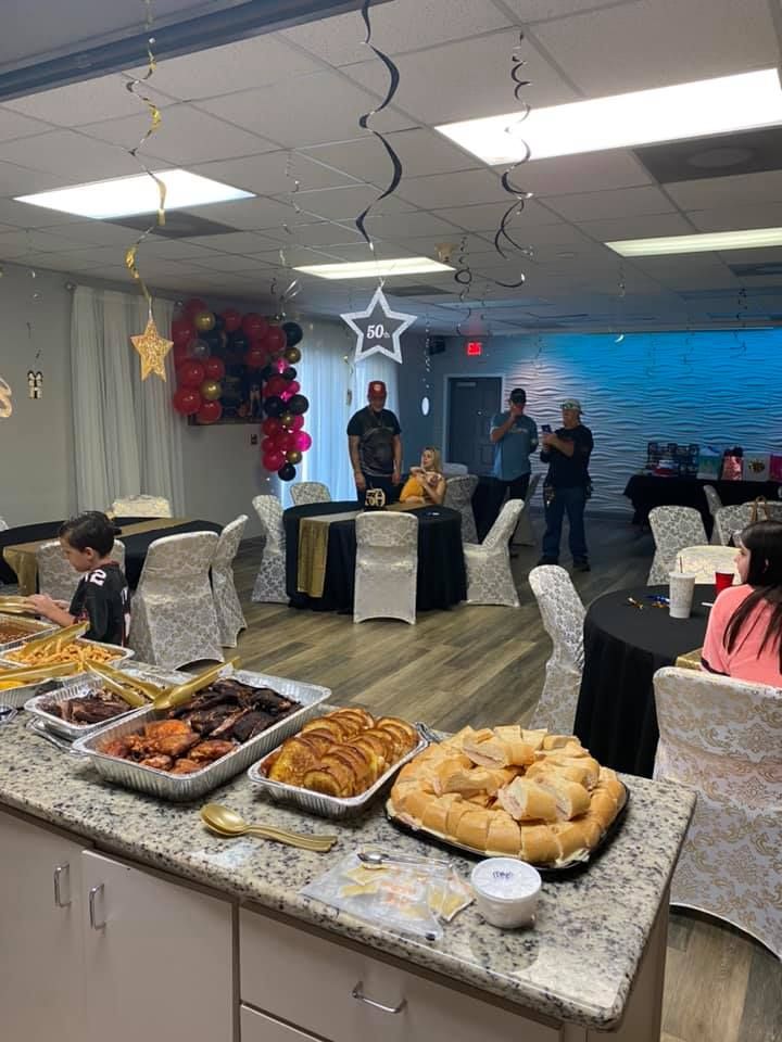 Party buffet with food on a counter, people in a room decorated with balloons and streamers.