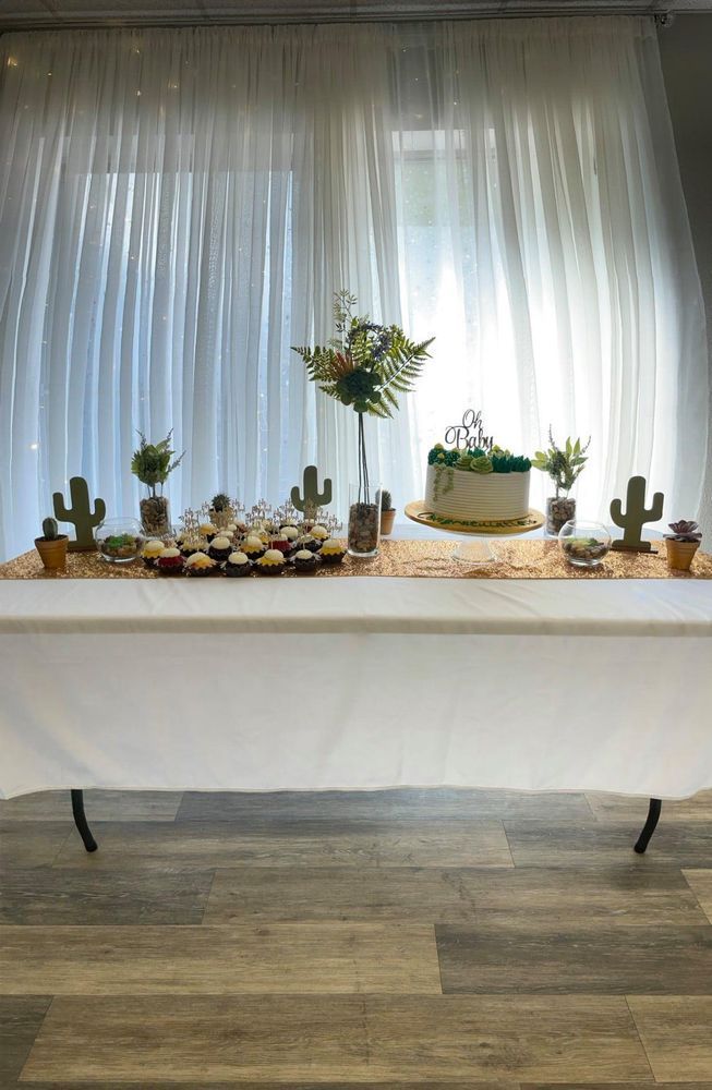 Desert-themed party table: cake, cupcakes, and cactus decorations on a white tablecloth with sheer curtains as a backdrop.