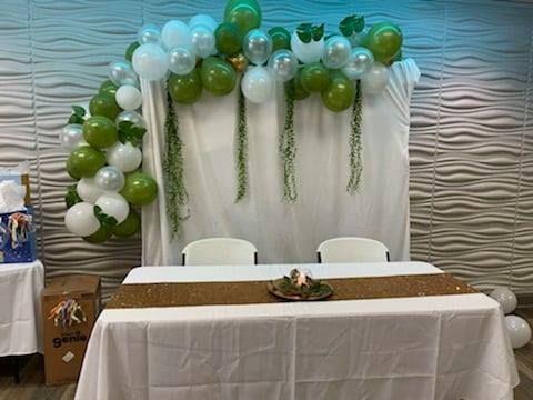 Decorated table with white tablecloth, green and white balloon arch, and hanging greenery.