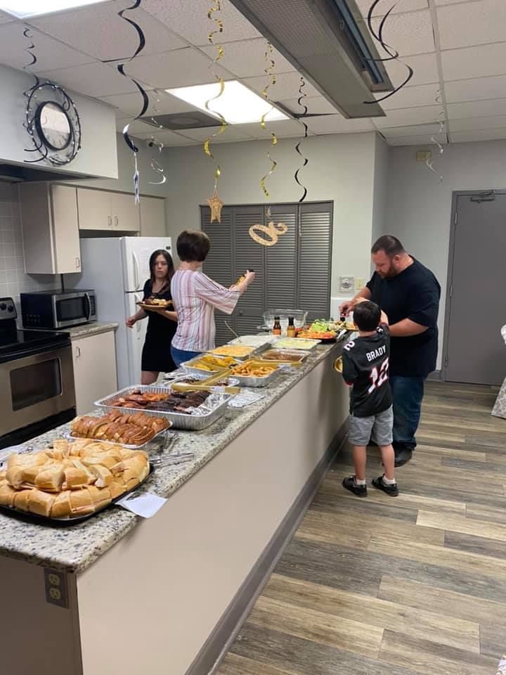 People at a kitchen counter with food. One person reaches toward food. Others stand nearby.