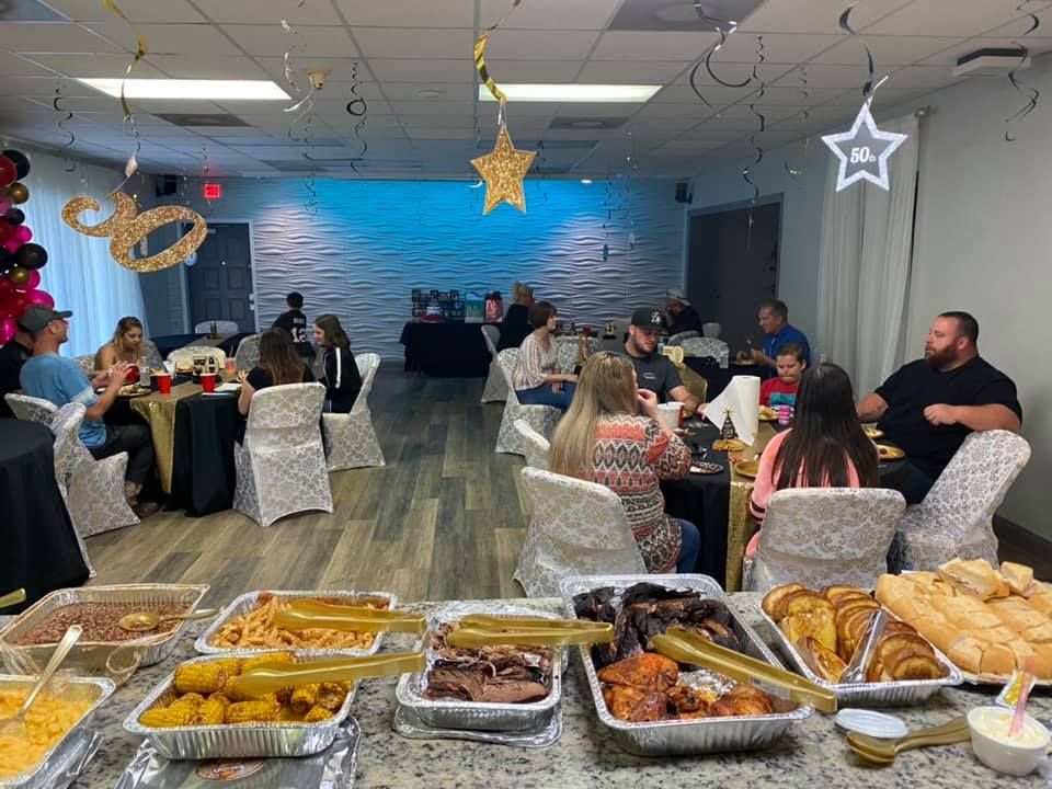 Buffet-style food setup at an event with people seated at tables, decorated with black, gold, and white.