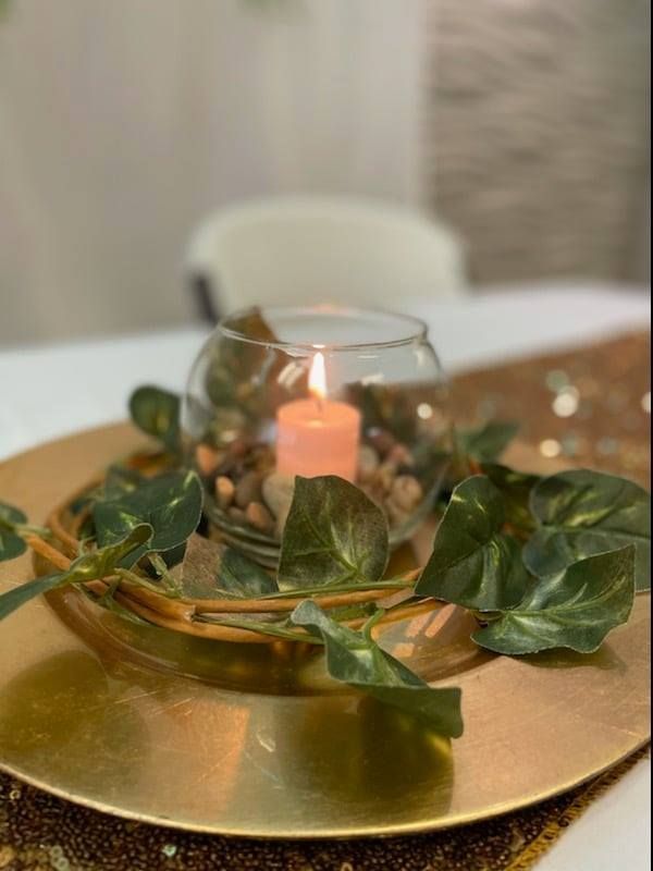 Candle in glass bowl with rocks and leaves on gold plate, table setting.