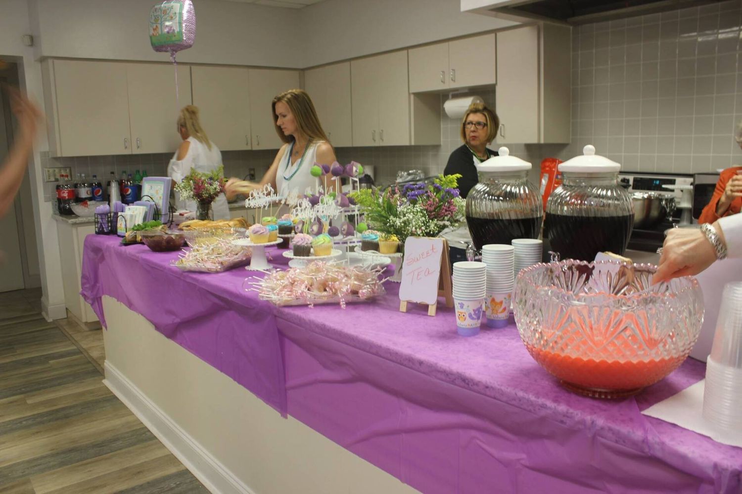 People at a party buffet table with food and drinks in a kitchen, purple tablecloth.