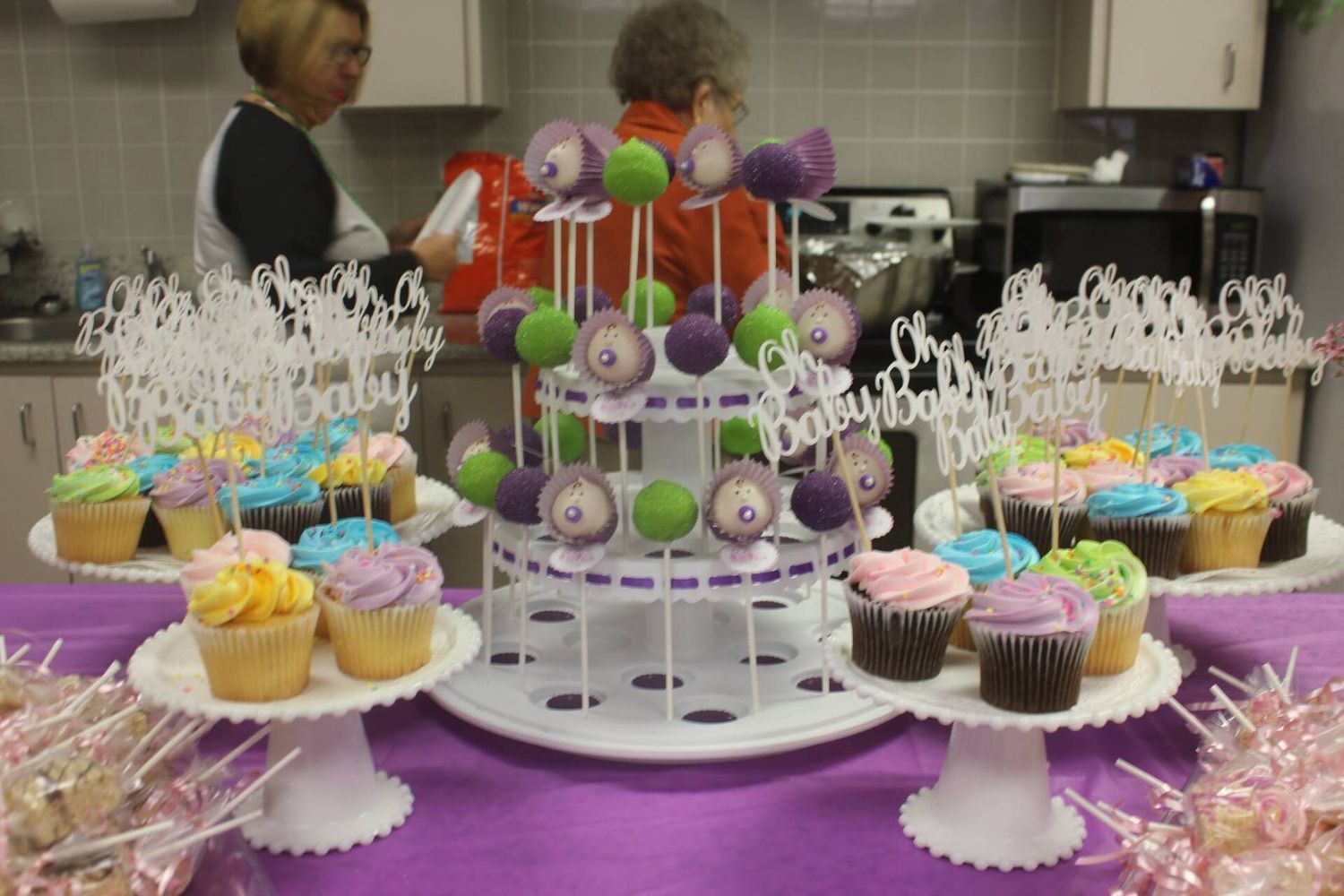 Dessert table with cupcakes, cake pops, and a tiered stand, purple and white decor, people in the background.