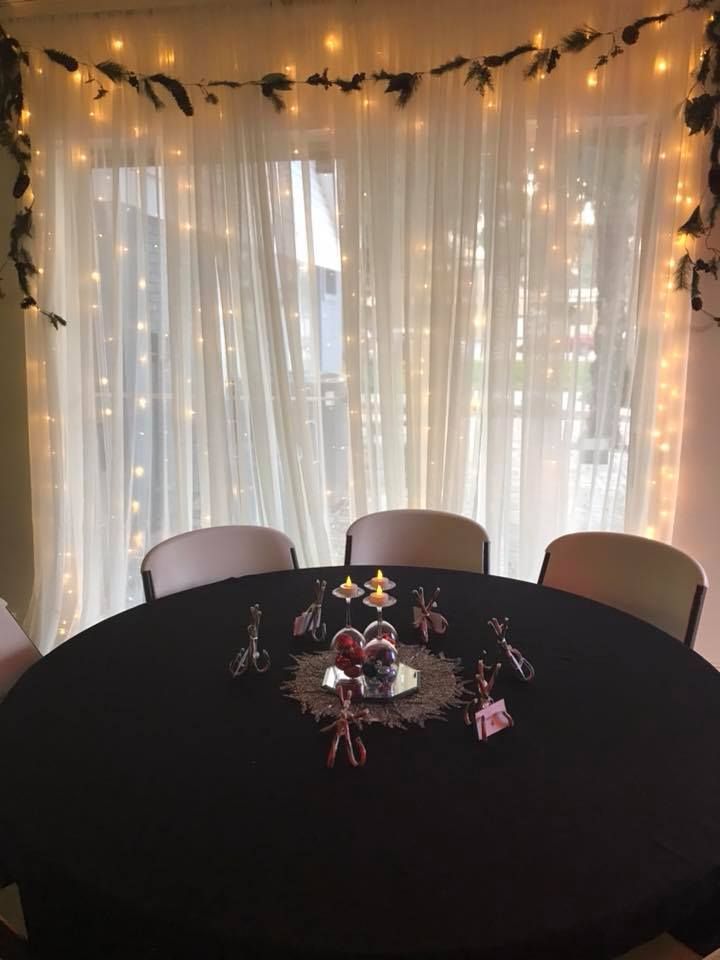 Round table with black cloth, three chairs, decorated window with lights and garland. Centerpiece.