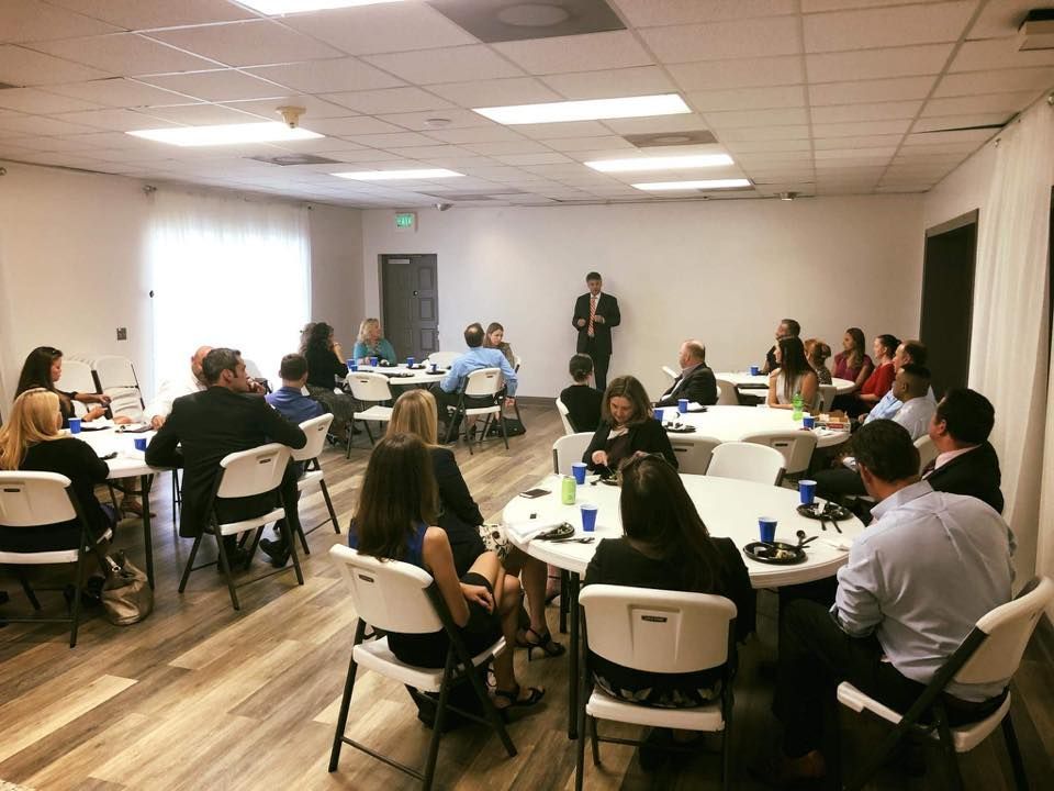 People in business attire at round tables in a conference room, one person speaking at the front.
