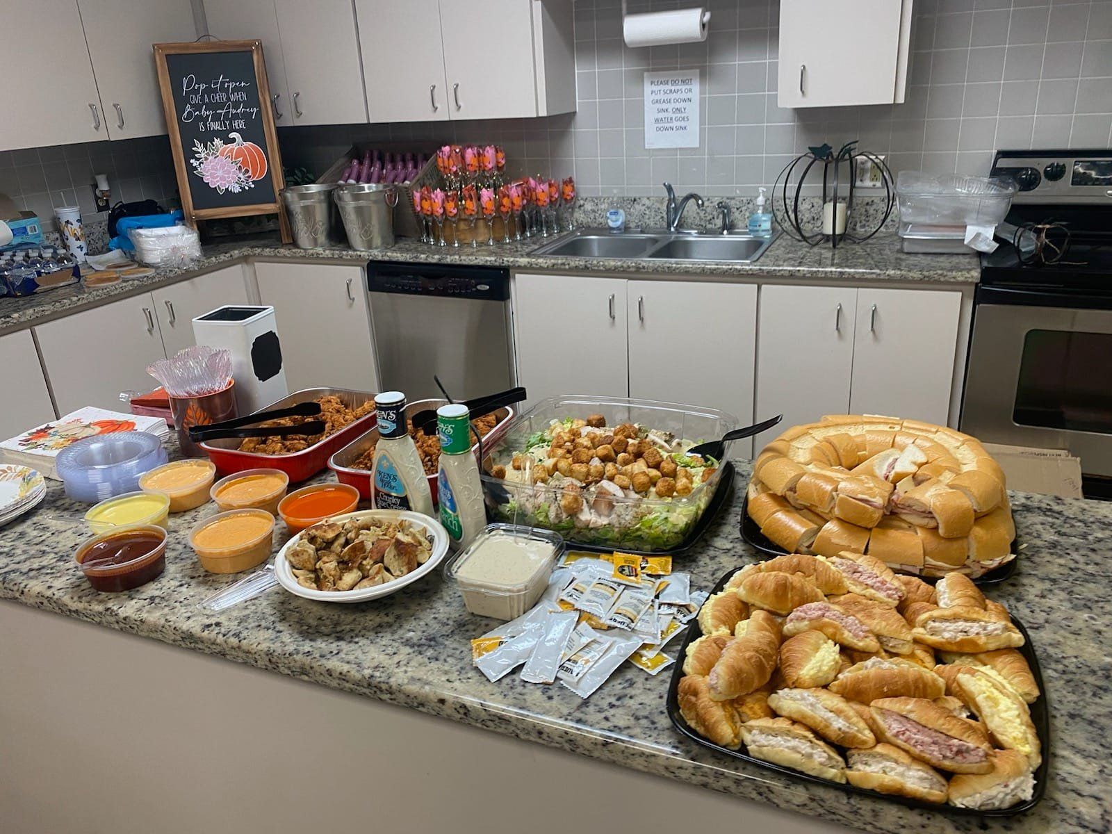 Kitchen counter with a spread of food: salads, dips, rolls, and other dishes. Cabinets and sink in background.