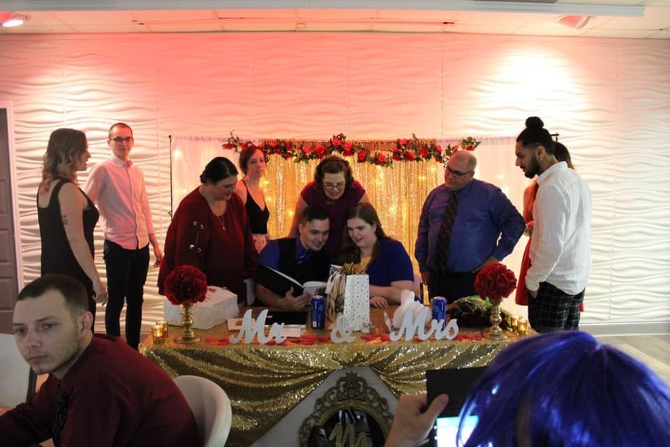 Wedding reception: Couple signing a marriage license at a decorated table; guests observe in front of a textured wall.