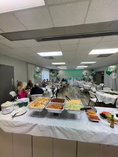 Buffet table with food; people seated at tables in a decorated room, balloons.