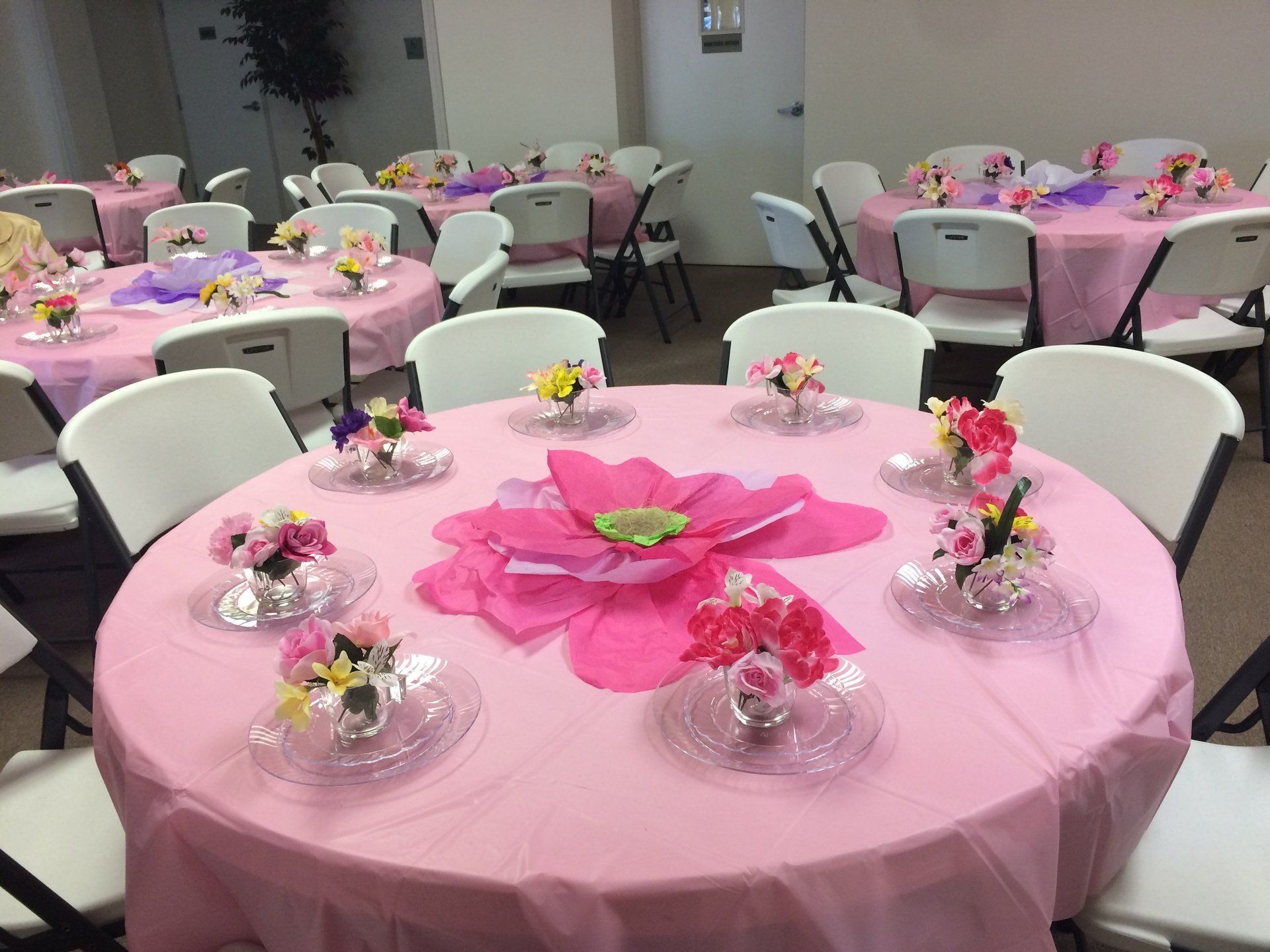 Round tables covered in pink cloths, set for a gathering. Floral centerpieces on each table.
