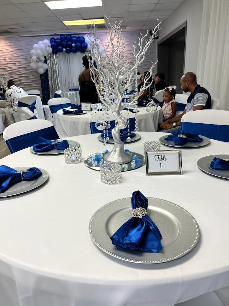 Elegant party table setting with silver chargers, blue napkins, and centerpiece; white tablecloth.