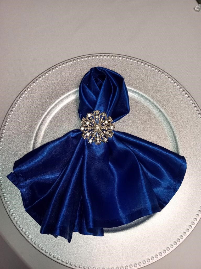 Blue napkin folded like a dress, held with a jewel, on a silver charger plate.