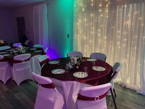 Round tables set for a party, with burgundy tablecloths and white chairs, illuminated by fairy lights.