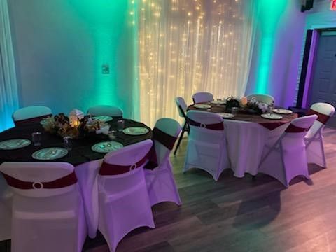 Round tables set for a gathering, white chair covers with maroon sashes, backdrop with lights, and colorful wall lighting.