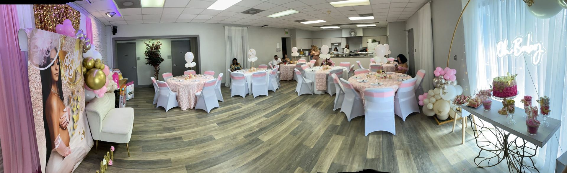 Room decorated for a party. Round tables with white covers and pink decor are set up. Light wood floors.