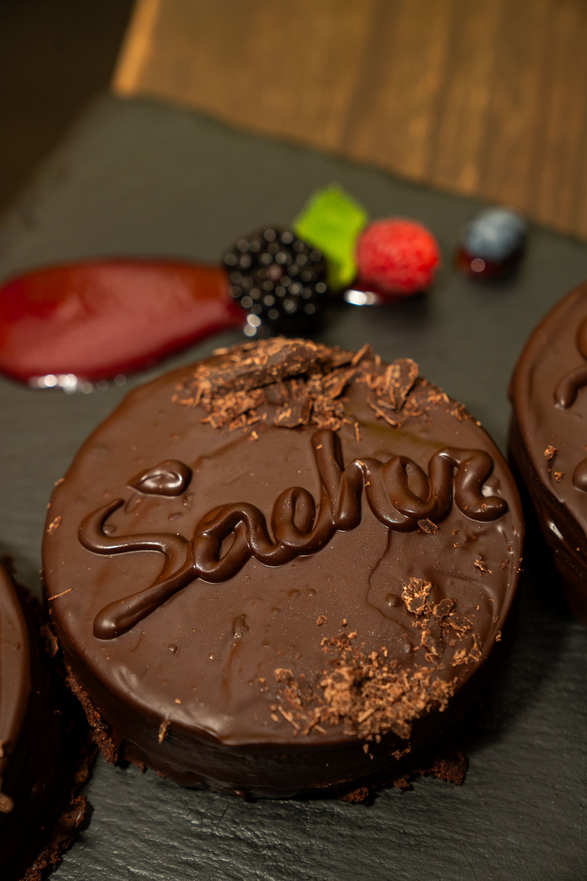 Dessert al cioccolato con glassa lucida e frutti di bosco su un piatto scuro, sormontato dalla scritta