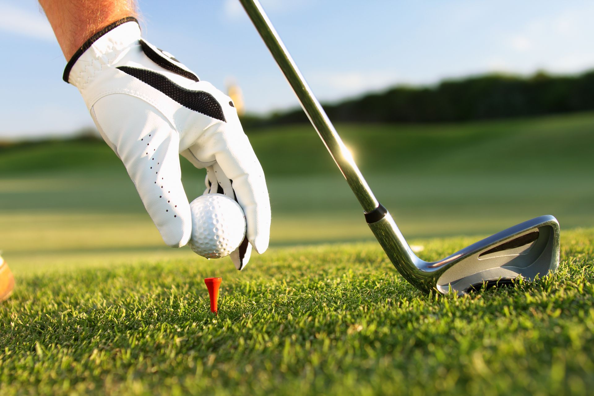 Golfer teeing up a golf ball on a tee with a golf club on the green, wearing a white golf glove, on a sunny day.