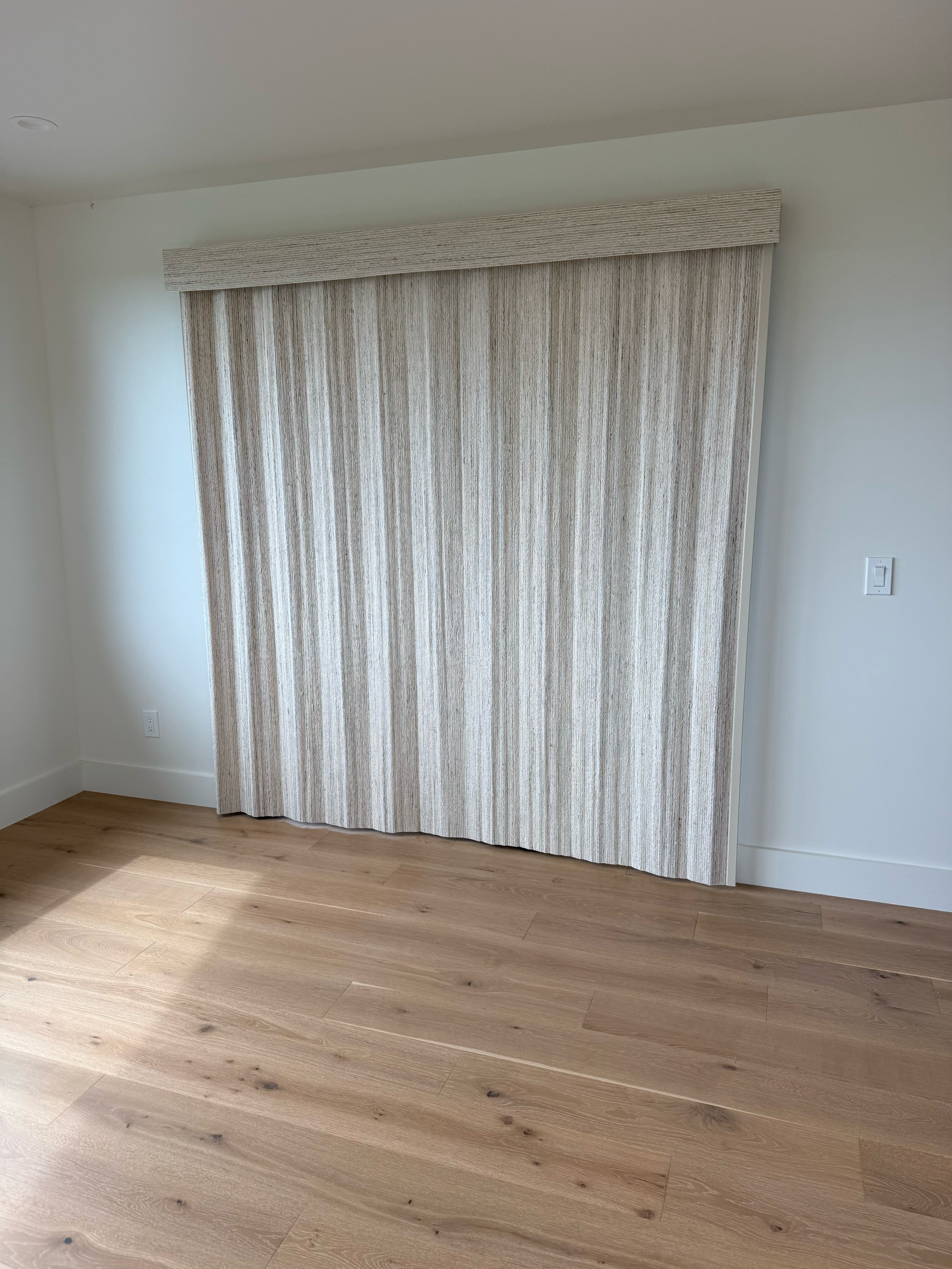 Empty room with beige curtains over a window and light wood flooring.