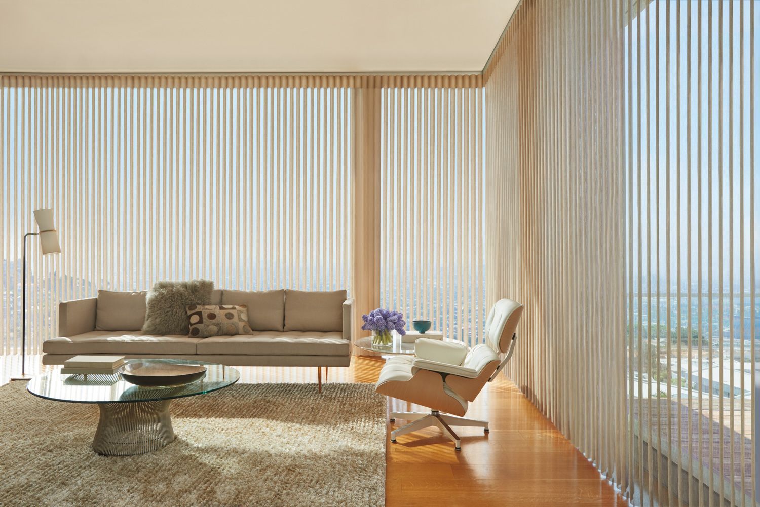 Modern living room with beige sofa, white chair, glass coffee table, and sheer floor-to-ceiling curtains.