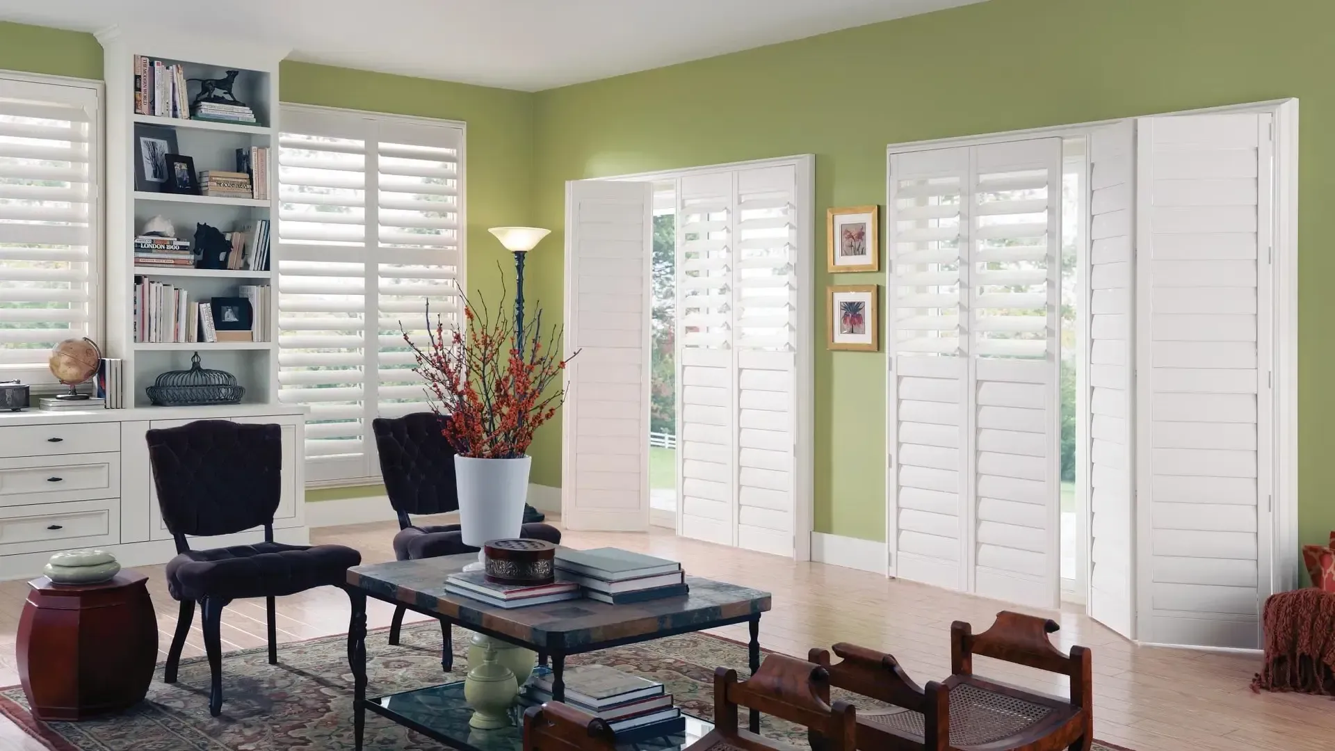 Bright living room with green walls, white shutters, dark furniture, and a table with flowers.