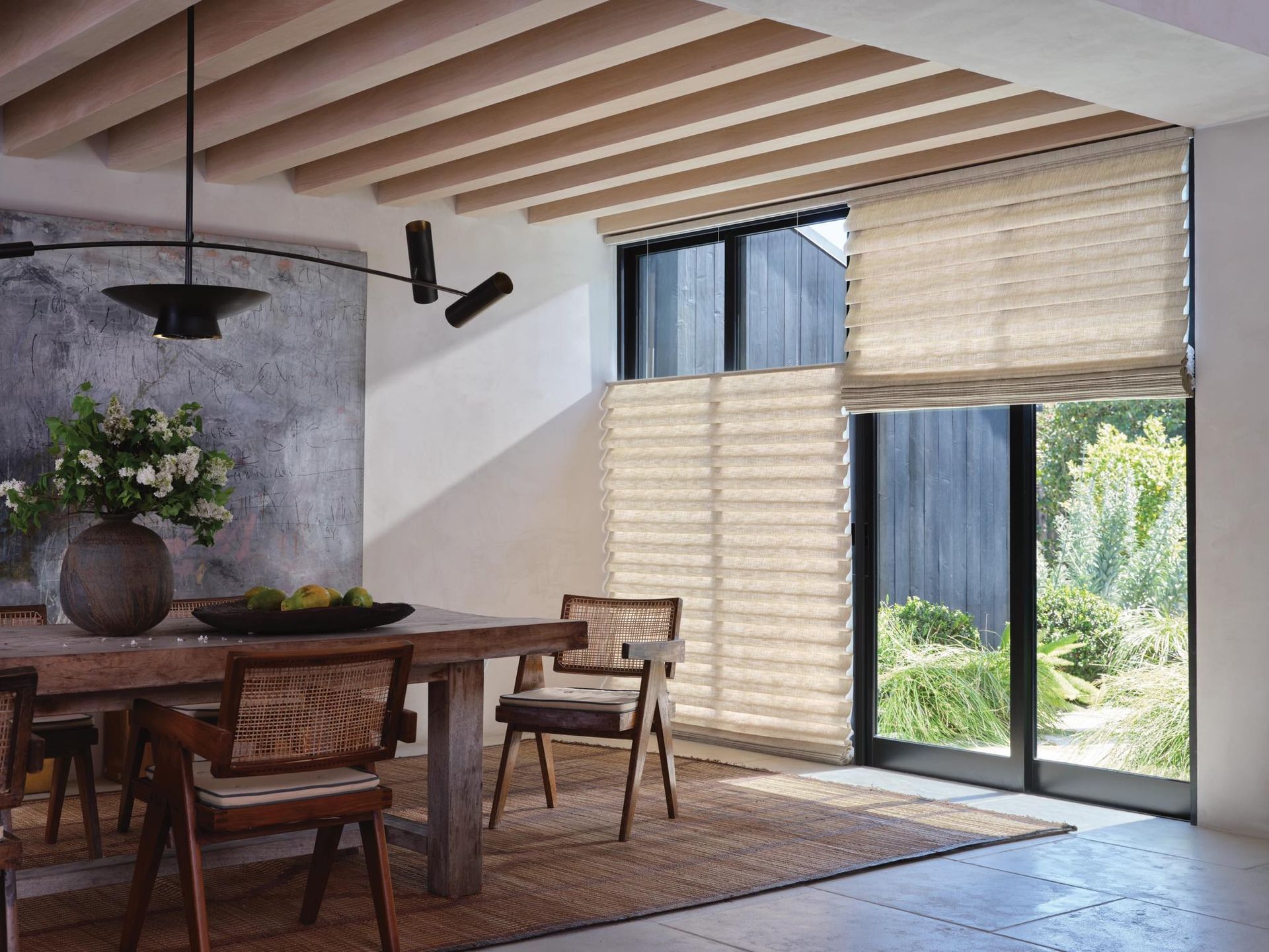Modern dining room with wooden table, woven chairs, skylight beams, and large window shades overlooking greenery