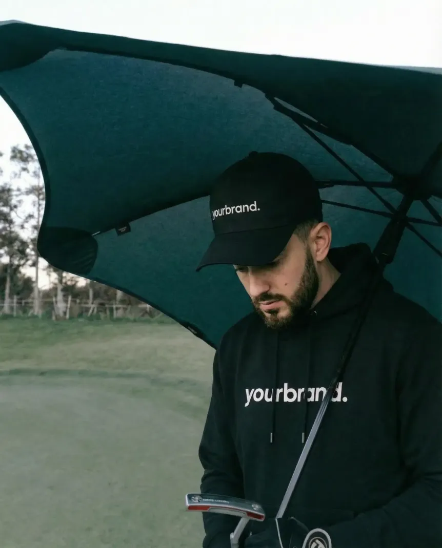 Model under a large teal umbrella, wearing a black cap and hoodie, looking down outdoors.