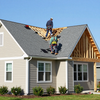 Two people in safety gear work on the exposed wooden frame of a grey shingled roof on a suburban house.