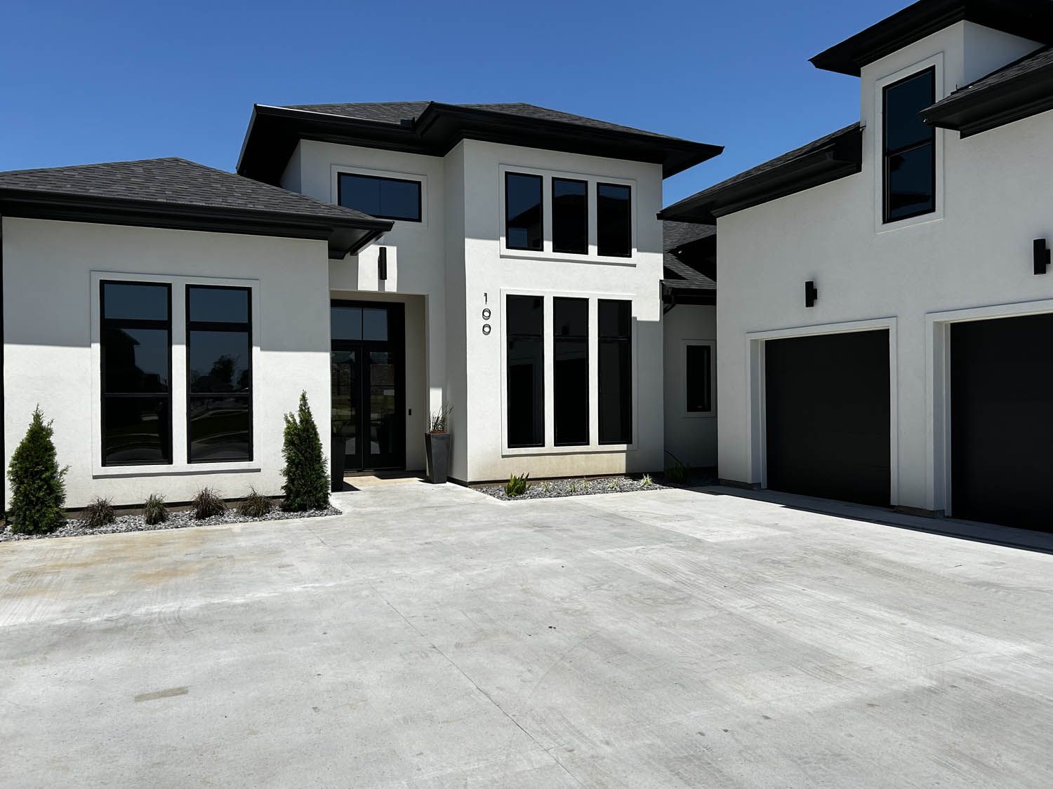Modern white house with dark trim and garage. Concrete driveway, large windows, and manicured landscaping.