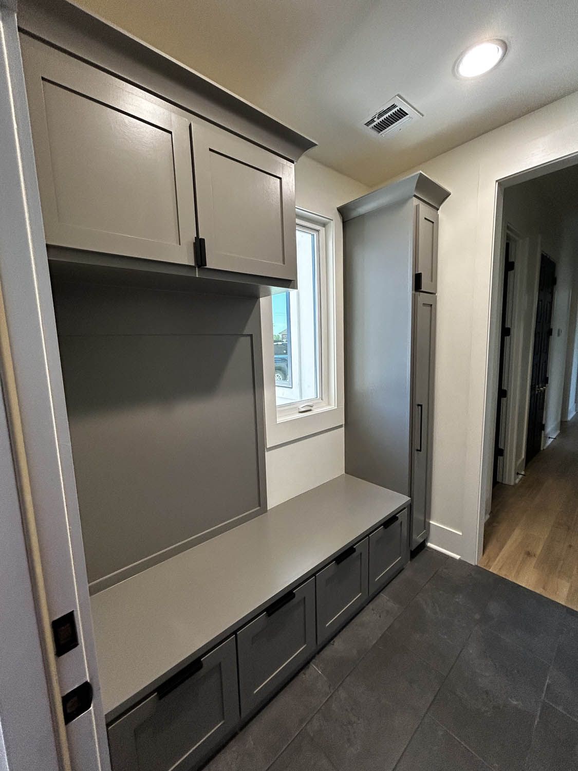 Gray built-in mudroom with storage cabinets, bench with drawers, and tall cabinet, next to a window and hallway.