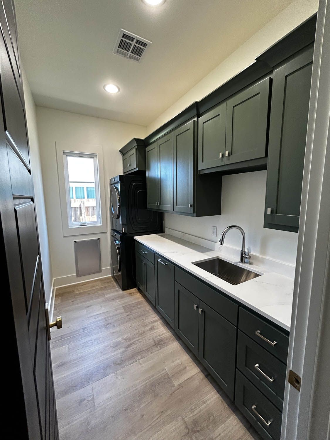 Dark green cabinets and white countertops in a laundry room with a stainless steel sink and window.